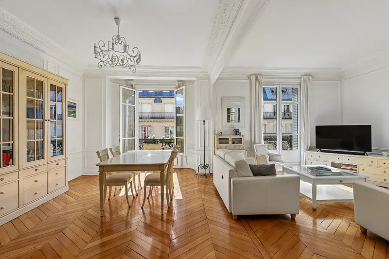 Bright living and dining room with herringbone wood floors, white furniture, a chandelier, and open balcony doors.