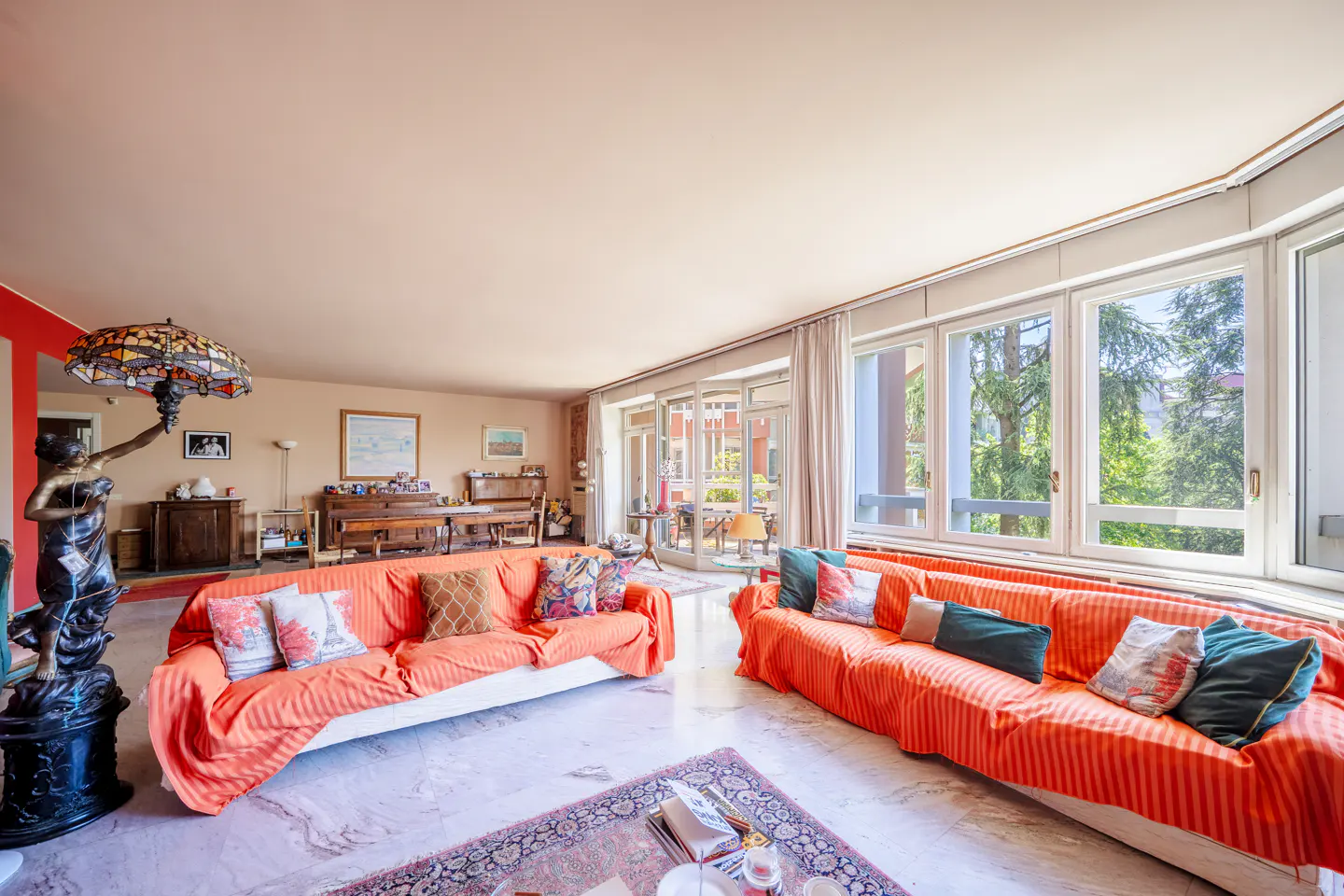 Bright living room with two orange sofas, marble floors, and large windows overlooking greenery. A statue with a lamp stands near a piano.