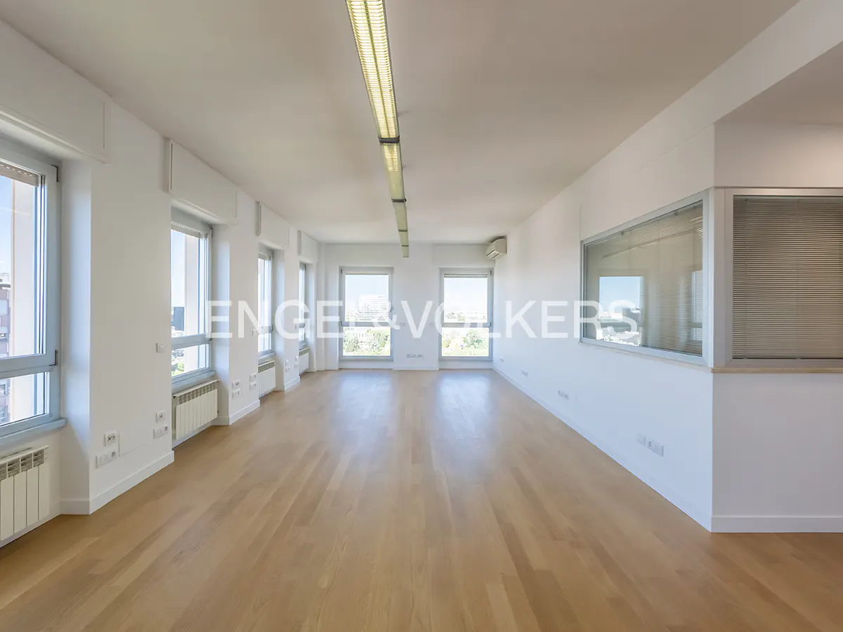 Bright, empty office space with hardwood floors, white walls, and large windows offering city views. Overhead fluorescent lighting.