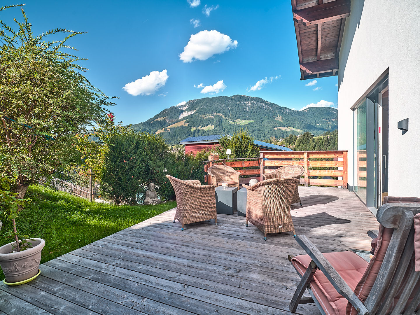 Sunny wooden deck with wicker patio furniture, overlooking a lush green lawn and scenic mountain range under a blue sky. [AI-generated text]