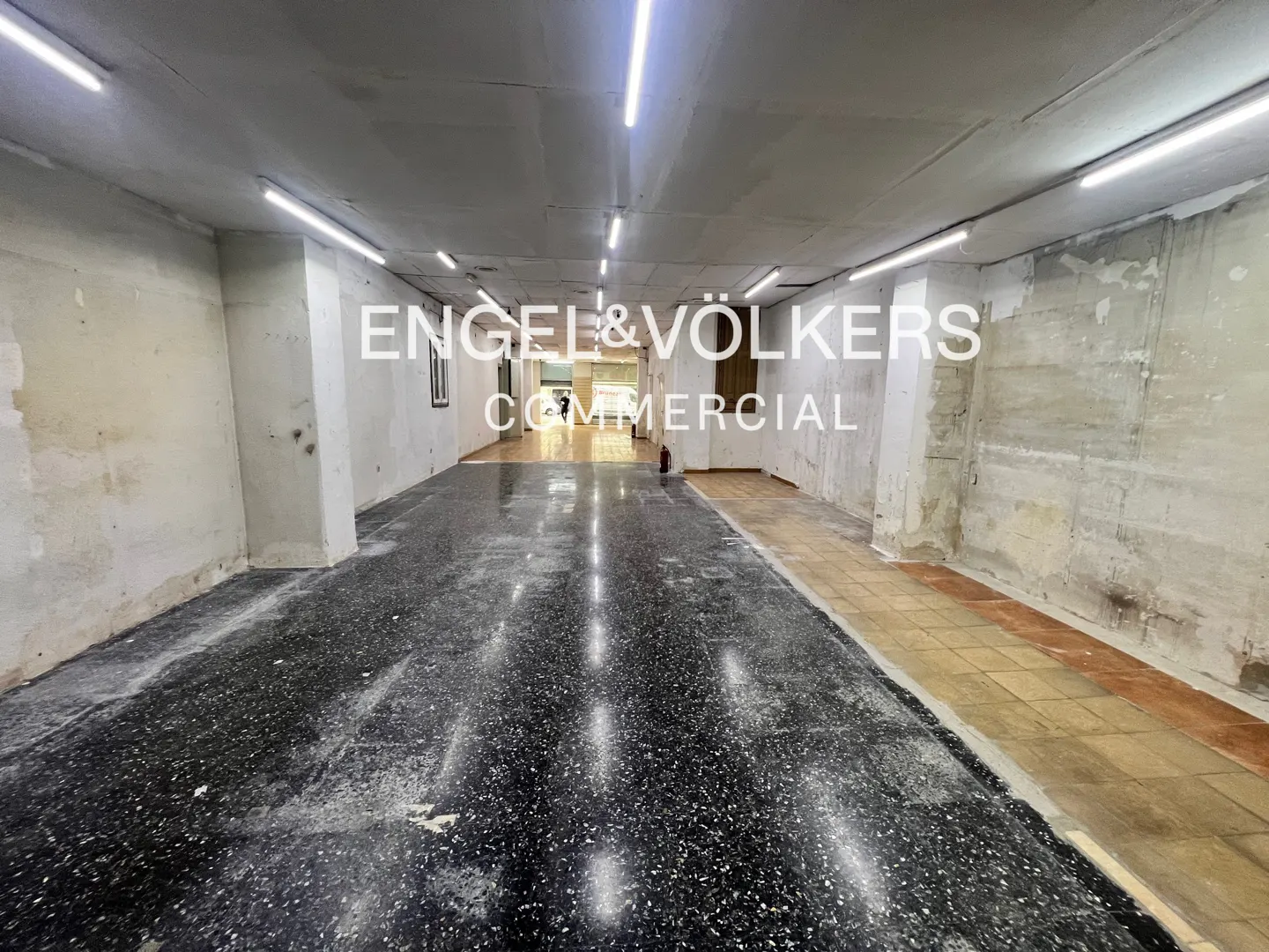 An empty commercial space with black speckled floors, white walls, and fluorescent lighting. The Engel & Völkers logo is superimposed on the image.