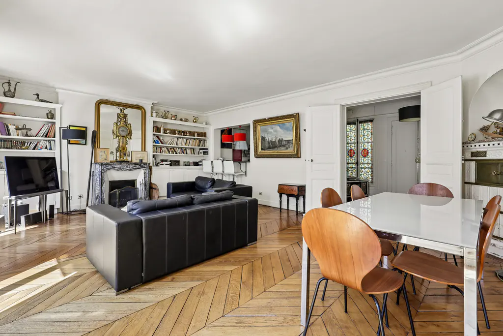 Bright living and dining area with herringbone wood floors. Black leather sofa, white bookshelves, and a white table with wood chairs.
