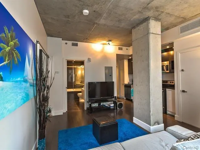 Living room with a blue rug, dark wood floors, and a concrete pillar. A beach scene mural is on the wall. The kitchen and bathroom are visible.