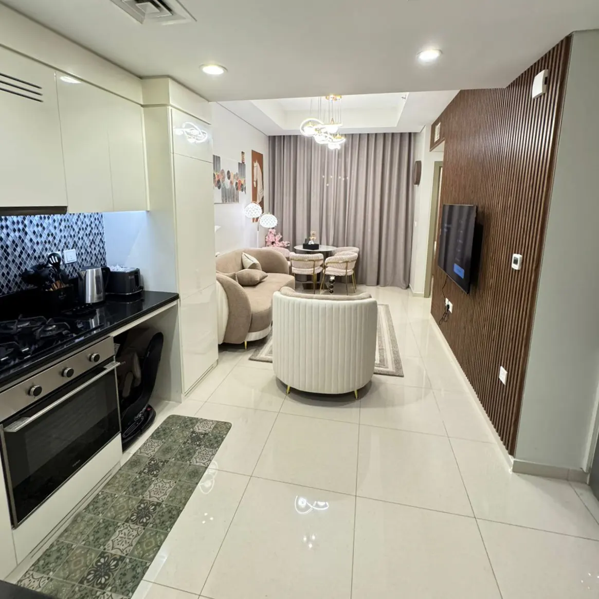 A modern apartment interior with a kitchen, living, and dining area. Cream-colored furniture, wood-paneled wall, and tile flooring.