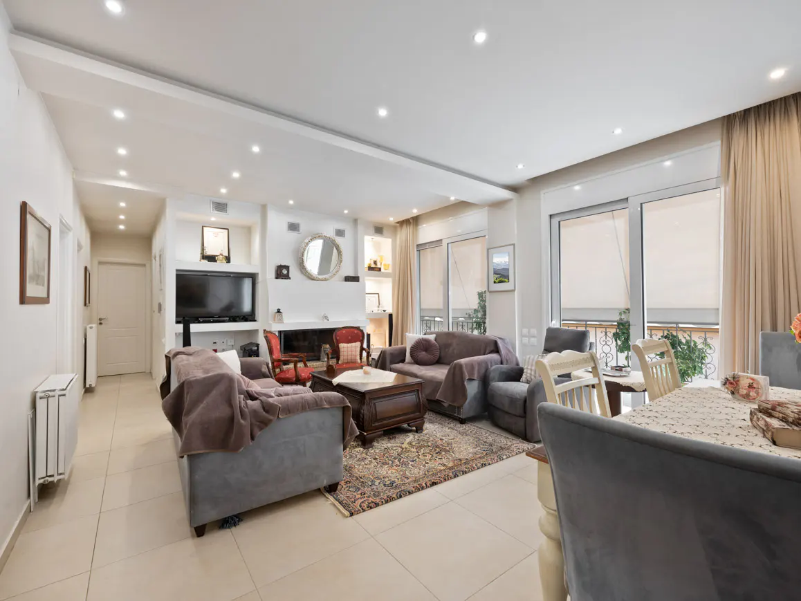 Bright living room with sofas, chairs, and a fireplace. Large windows with beige curtains let in natural light. A dining table is visible.