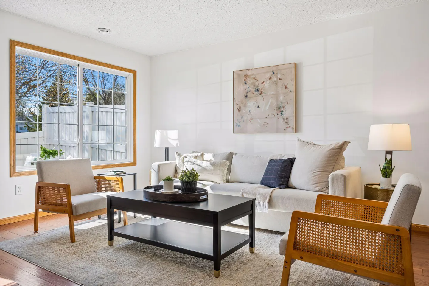 Bright living room with white walls, a beige sofa, and two cane-backed chairs. A black coffee table sits on a neutral rug. Large window with a view.
