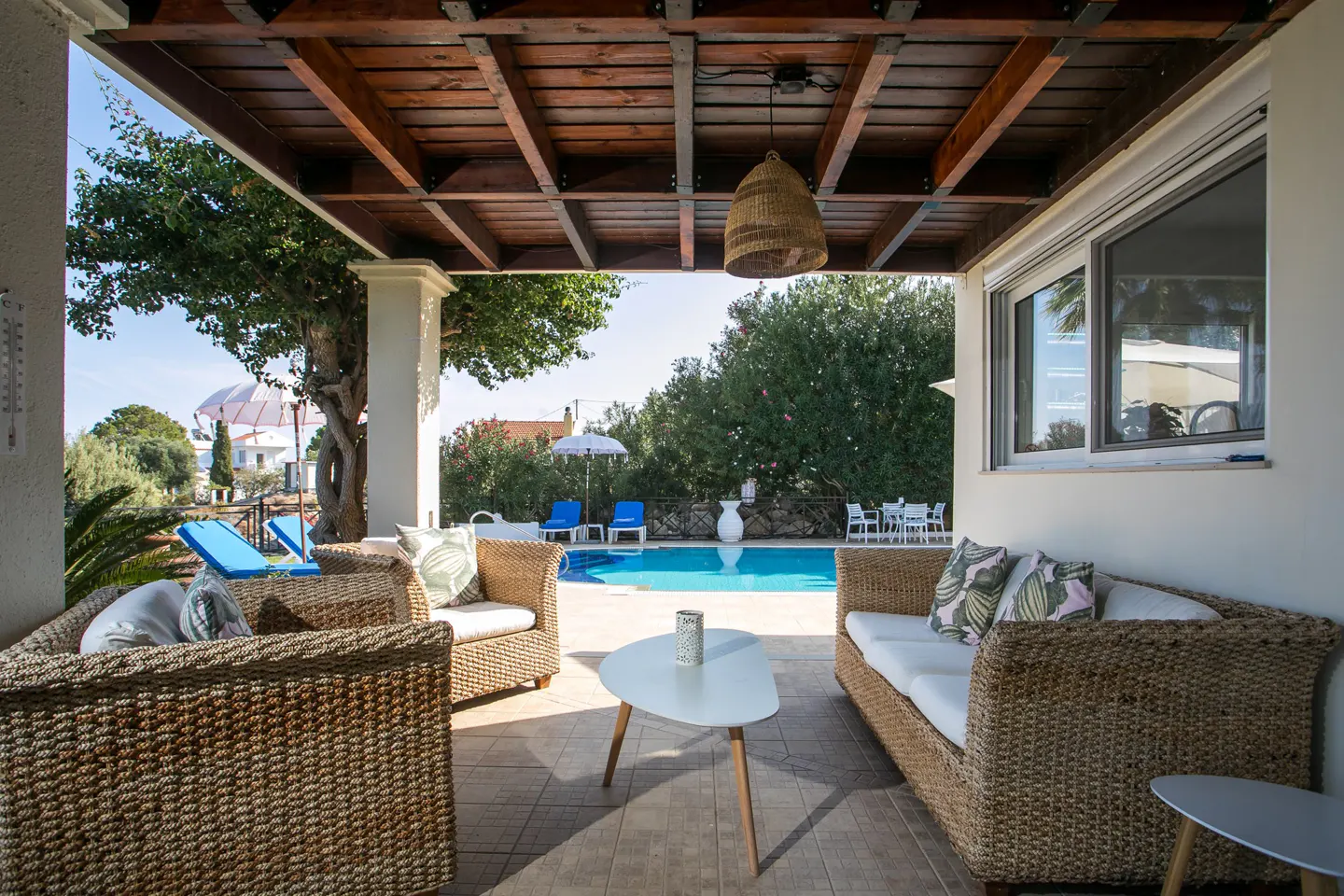 Covered patio with wicker furniture overlooks a blue pool and lush greenery. A wooden ceiling and hanging light fixture add warmth.