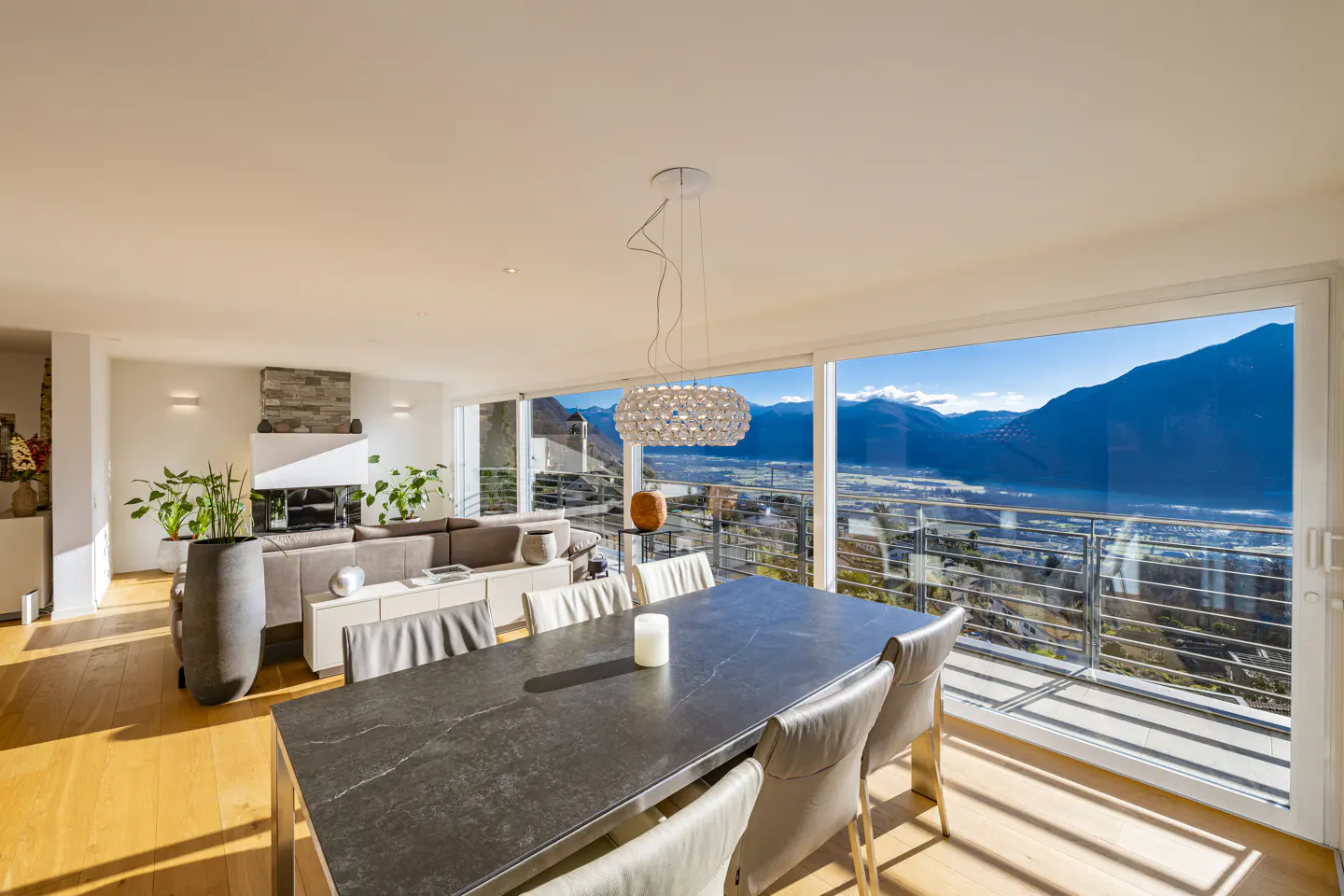 Bright, modern living room with a dark dining table, light chairs, and a mountain view through large windows.