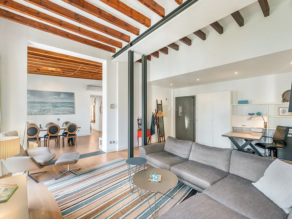 Bright, modern living room with exposed wood beams, gray sofa, striped rug, and dining area visible through an archway.