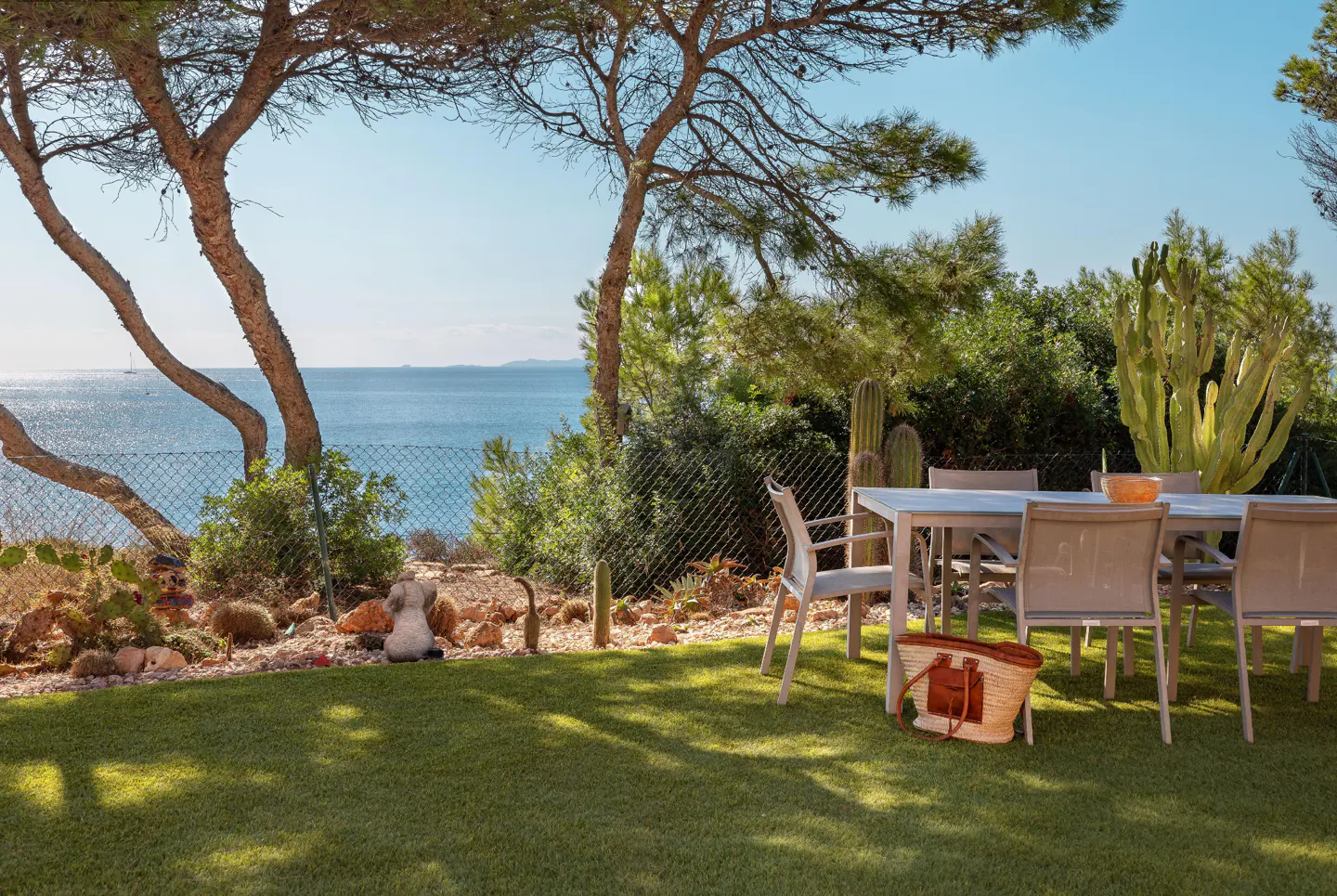 Outdoor patio with table, chairs, and straw bag on green lawn. Ocean view with trees and cacti in the background.