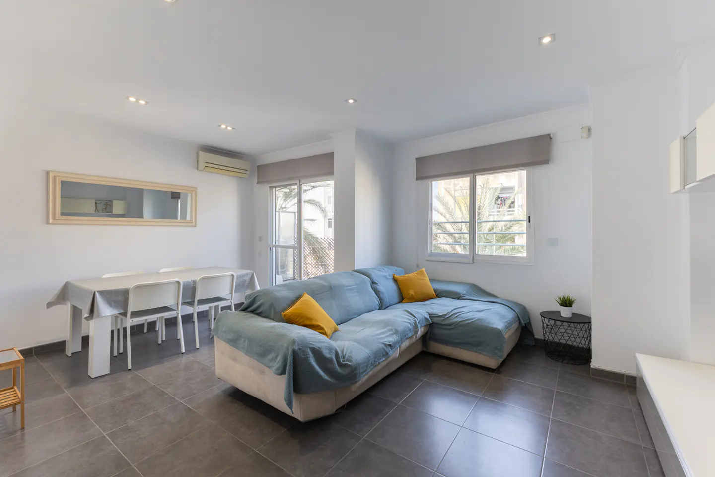 Bright living room with a blue sectional sofa, yellow pillows, dining table with white chairs, and gray tile flooring.