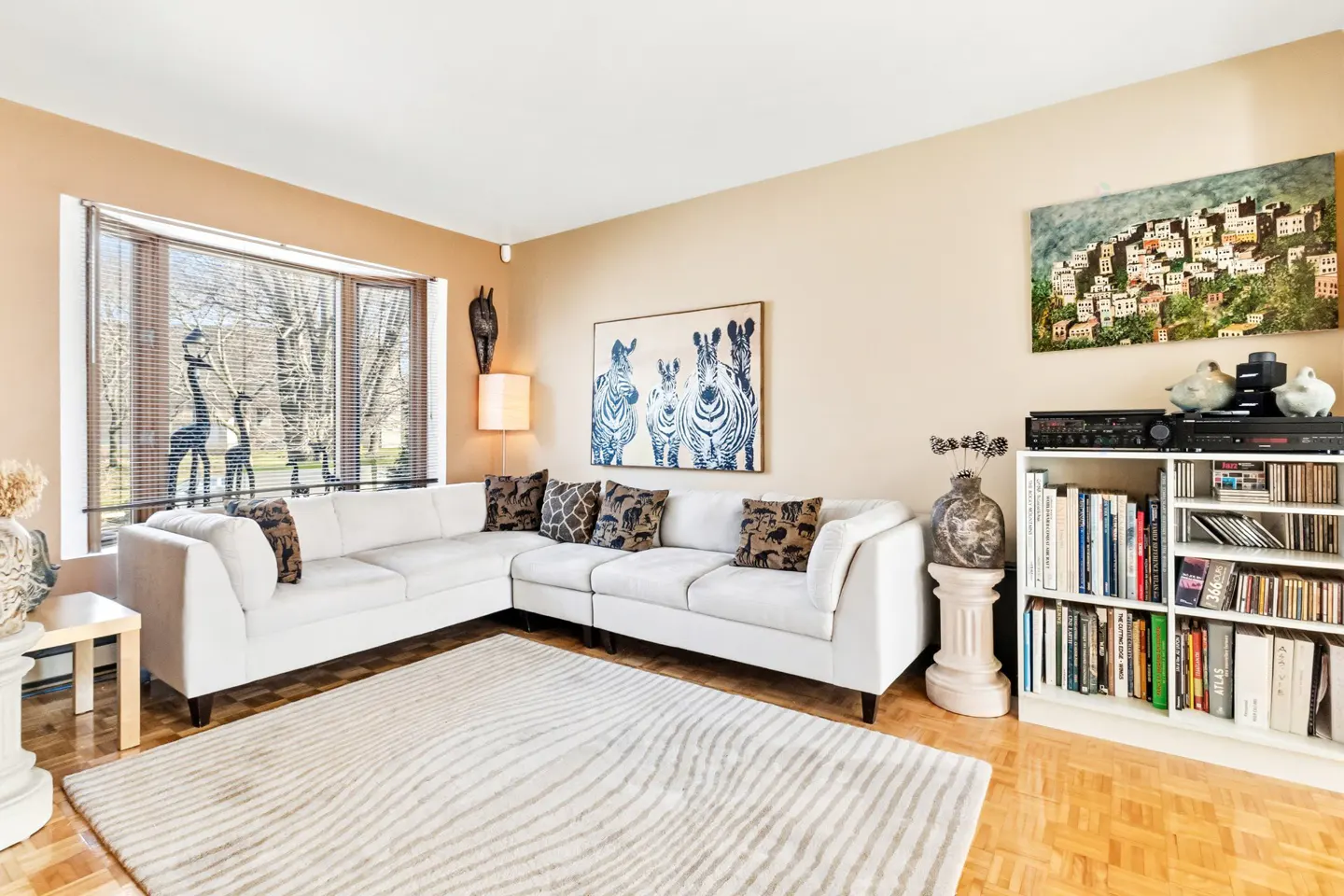 Living room with a white sectional sofa, zebra art, and a bookshelf filled with books and CDs. A large window offers a view of trees.