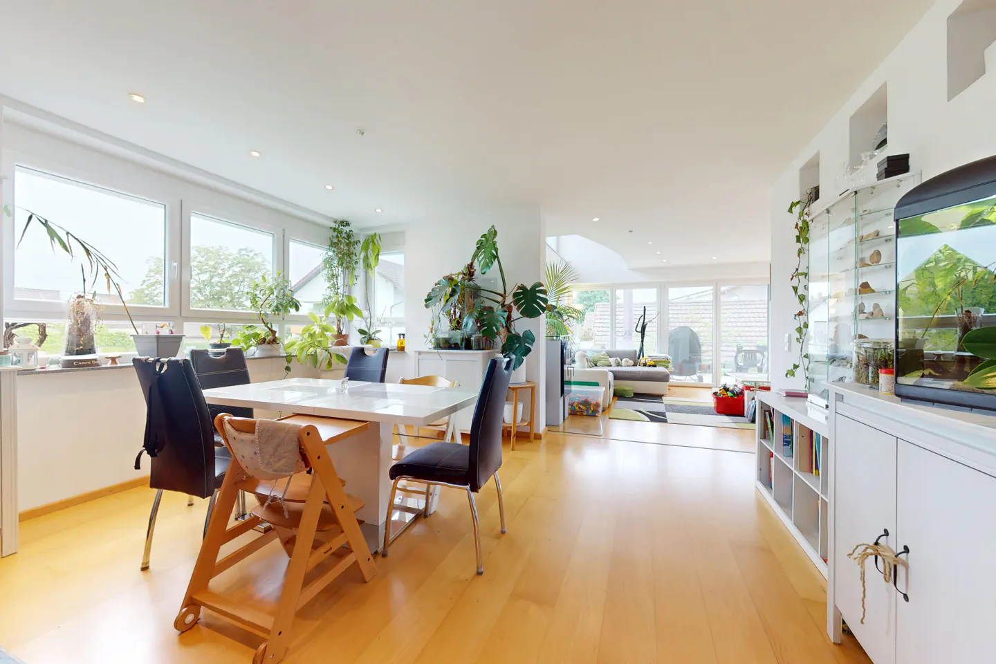 Bright, open-concept dining area with a white table, black chairs, and a wooden high chair. Large windows, plants, and an aquarium add natural elements.