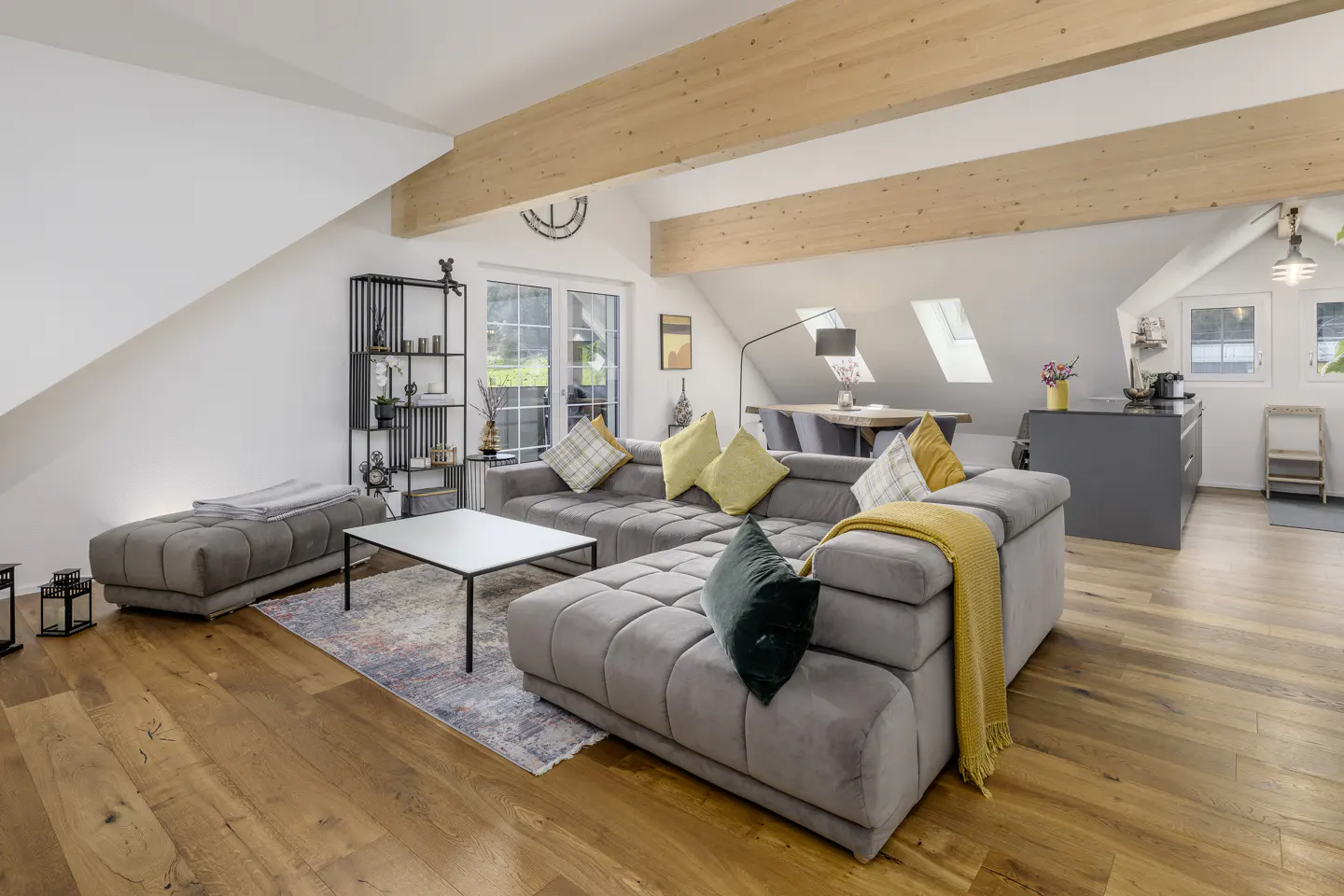 Bright living room with wood floors, exposed beams, and a gray sectional sofa with yellow and green pillows. A modern kitchen is visible in the background.
