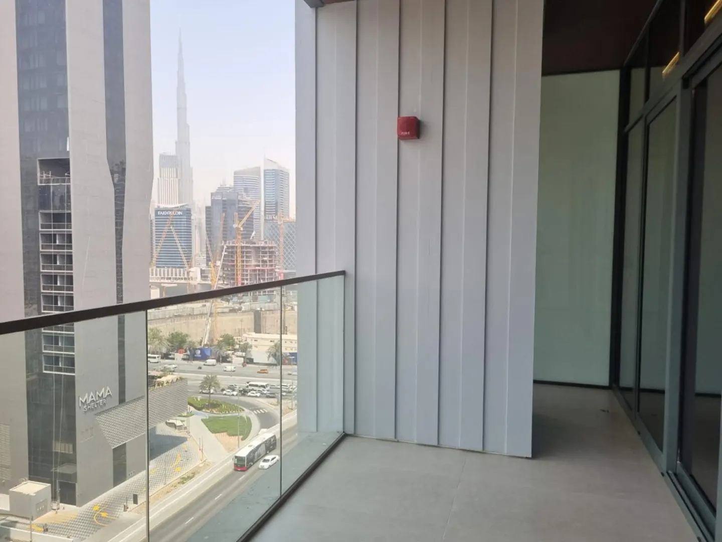Balcony view of Dubai skyline with Burj Khalifa. Glass railing, grey walls, and a red fire alarm box are visible.