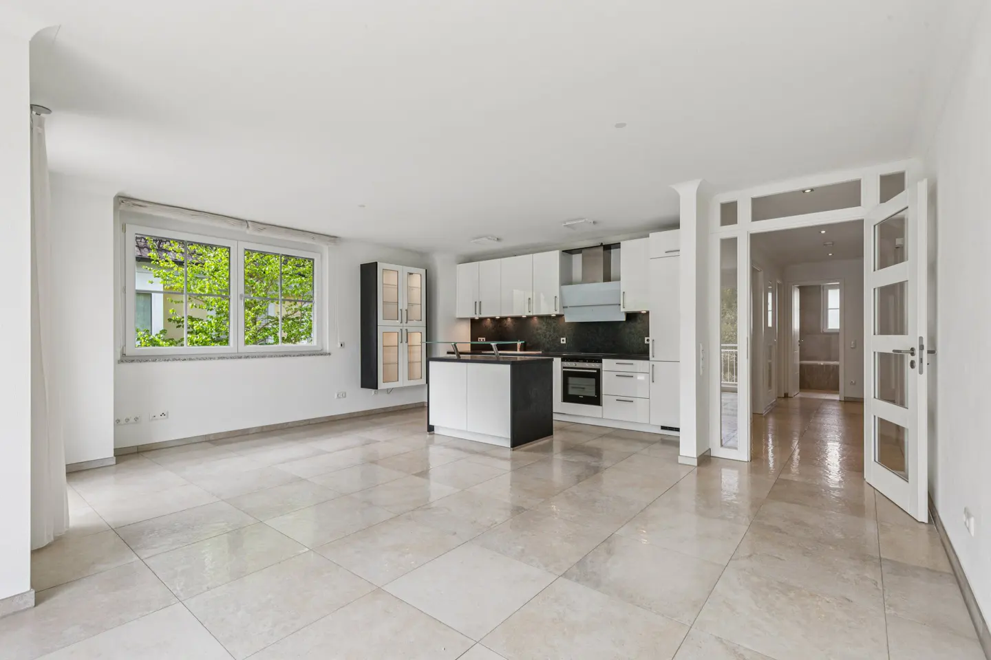 Bright, open-concept kitchen and living area with white cabinets, island, and tile flooring. A window shows green trees outside.