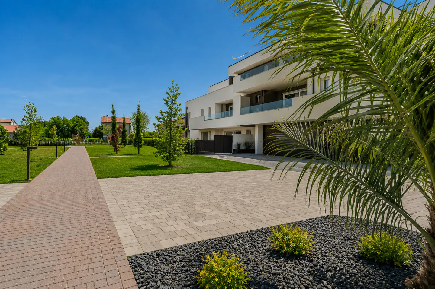 Modern white building with balconies, a paved driveway, and a palm tree on a sunny day.