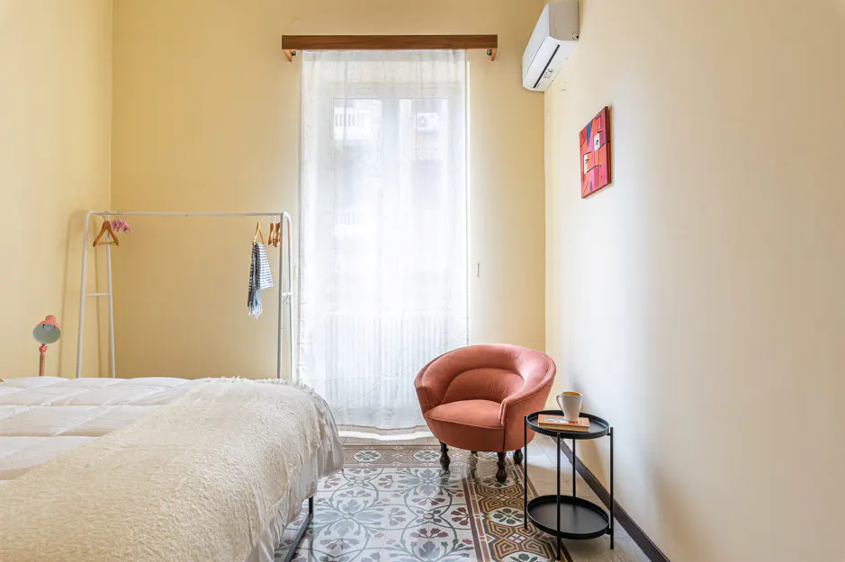 Bedroom with a white bed, clothes rack, and salmon-colored chair. A small black table sits next to the chair. The walls are light yellow.