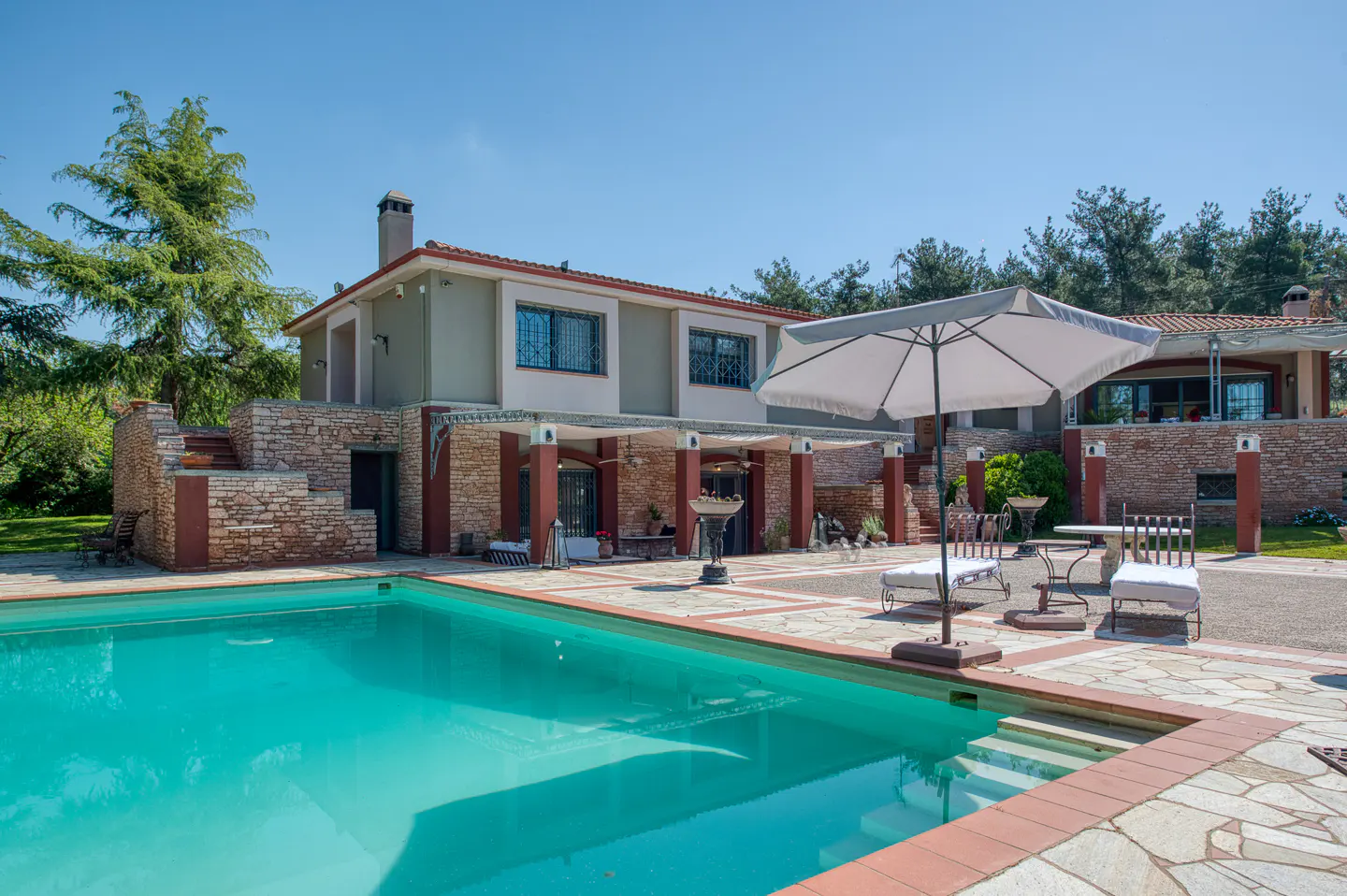 Exterior view of a two-story house with a pool, patio, and white umbrella. The house is stone and stucco with red trim. The pool is turquoise.