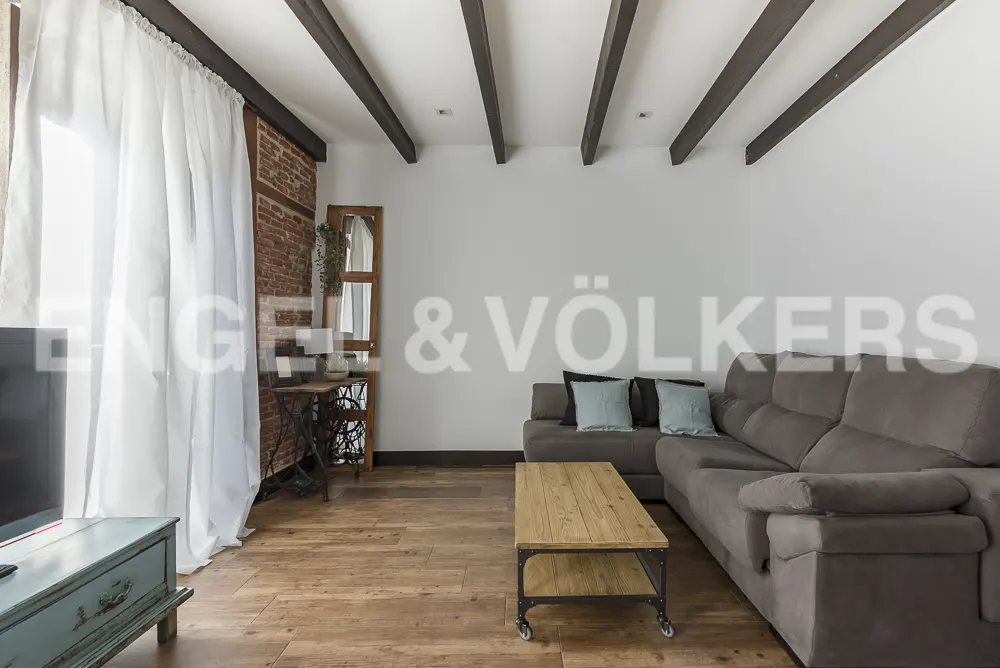 Living room with exposed brick, wood beams, and hardwood floors. A gray sectional sofa and wood coffee table are in the center. White curtains hang by the window.