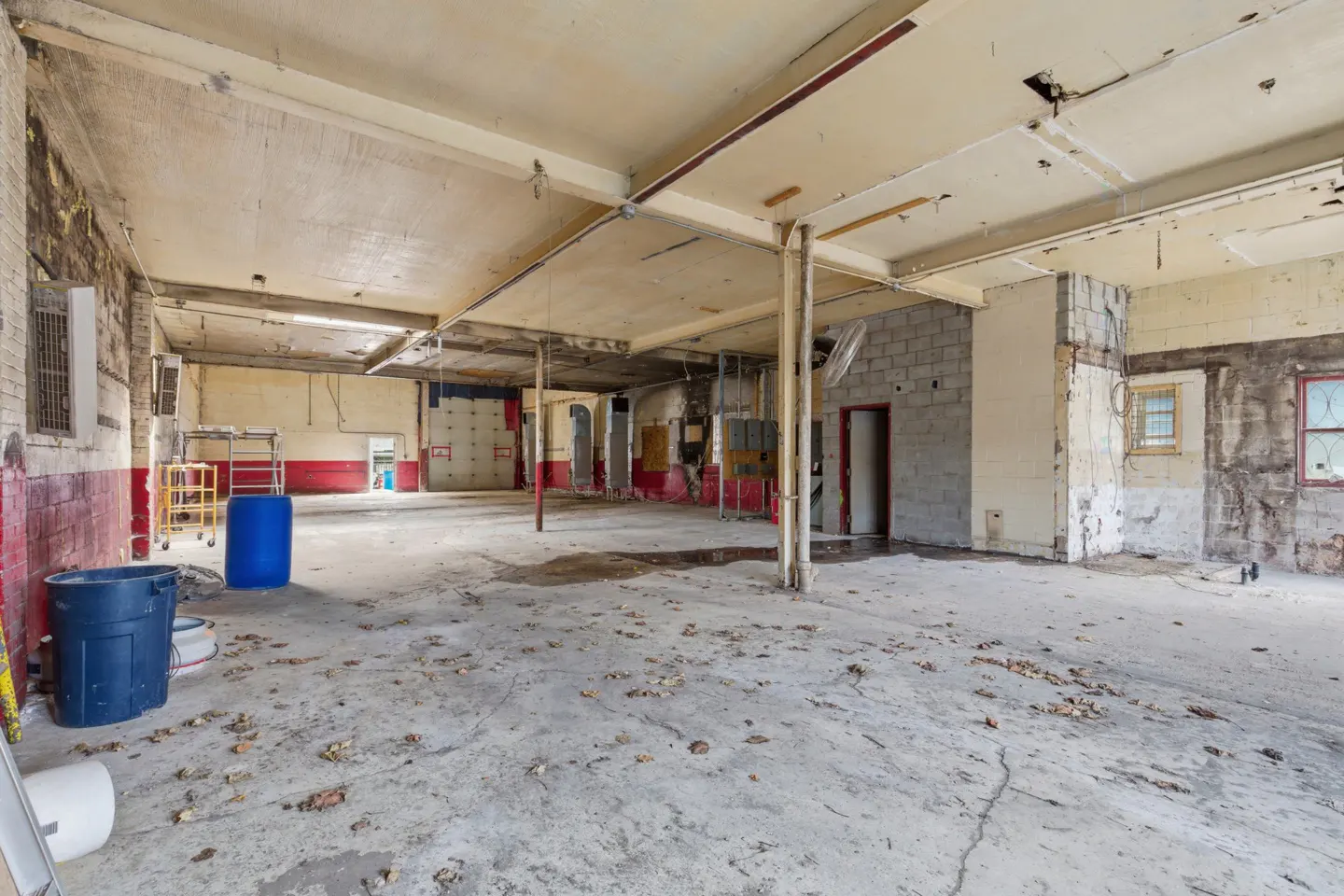 Large, empty commercial space with concrete floors, exposed ceiling, and red and white brick walls. Blue barrels and a yellow cart are visible.