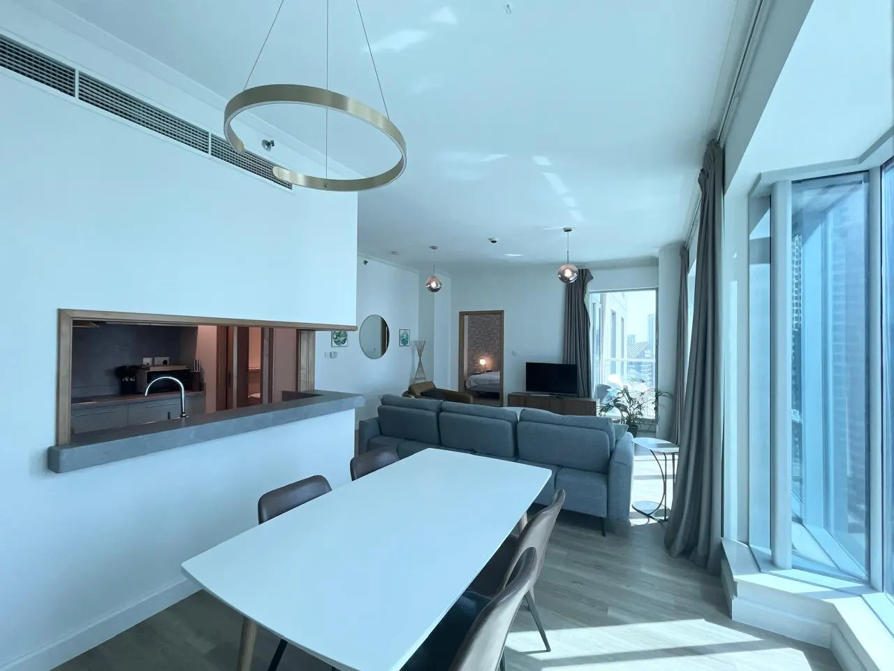 Bright, modern apartment interior with a white dining table, gray sofa, and large windows. A circular light fixture hangs above.