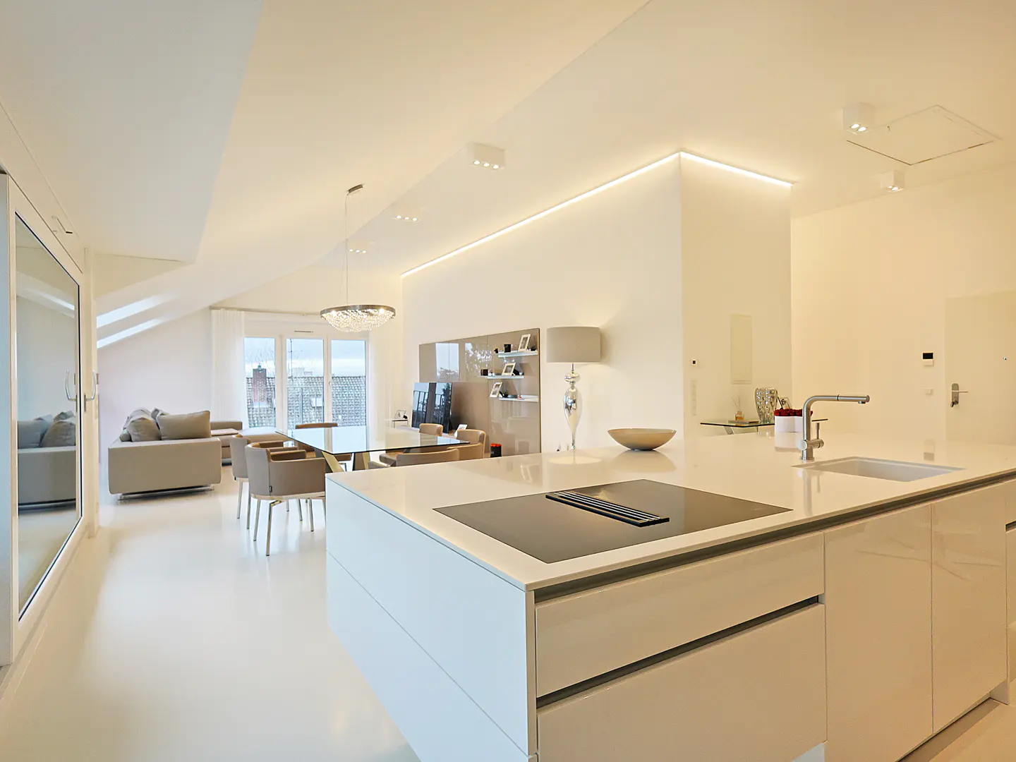 Bright, open-concept apartment with white walls and floors. A kitchen island with a cooktop is in the foreground, with a dining table and living room visible in the background.