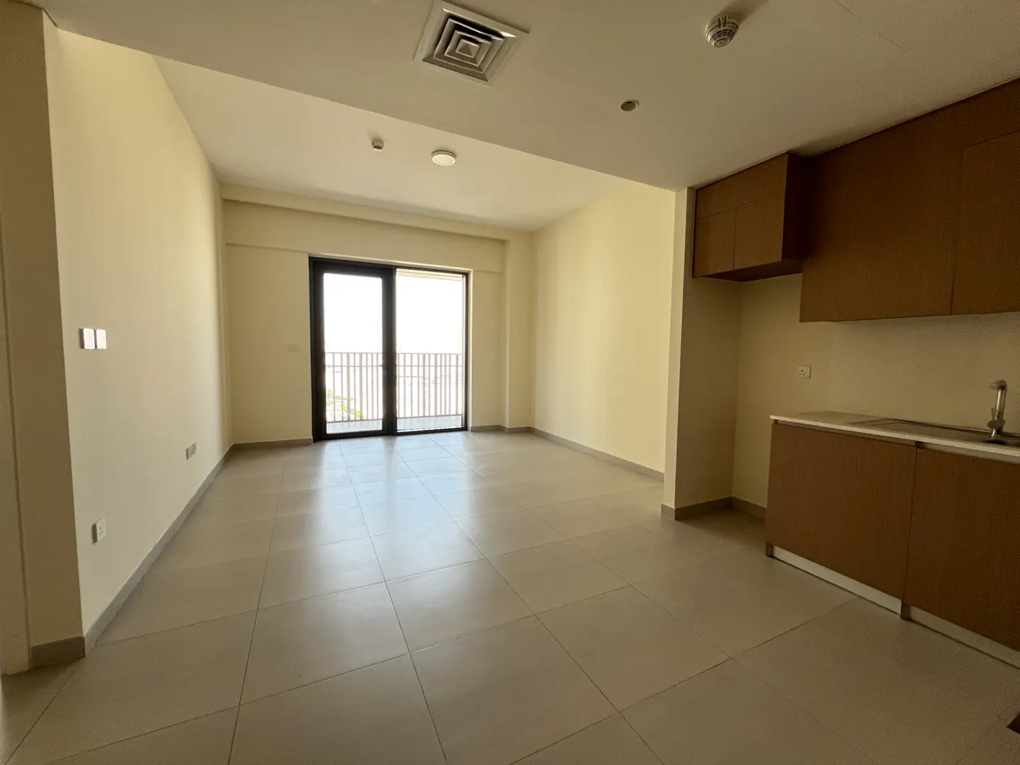 Empty apartment living area with light tile floors, beige walls, and a small kitchen with brown cabinets.