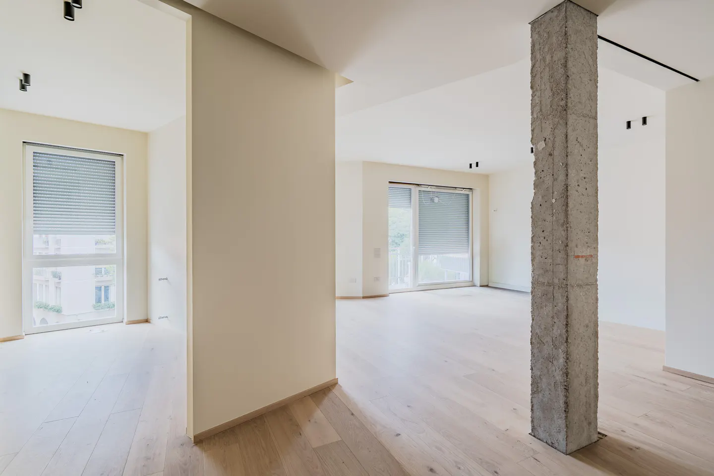 Minimalist interior with light wood floors, cream walls, large windows with blinds, and an exposed concrete support pillar.