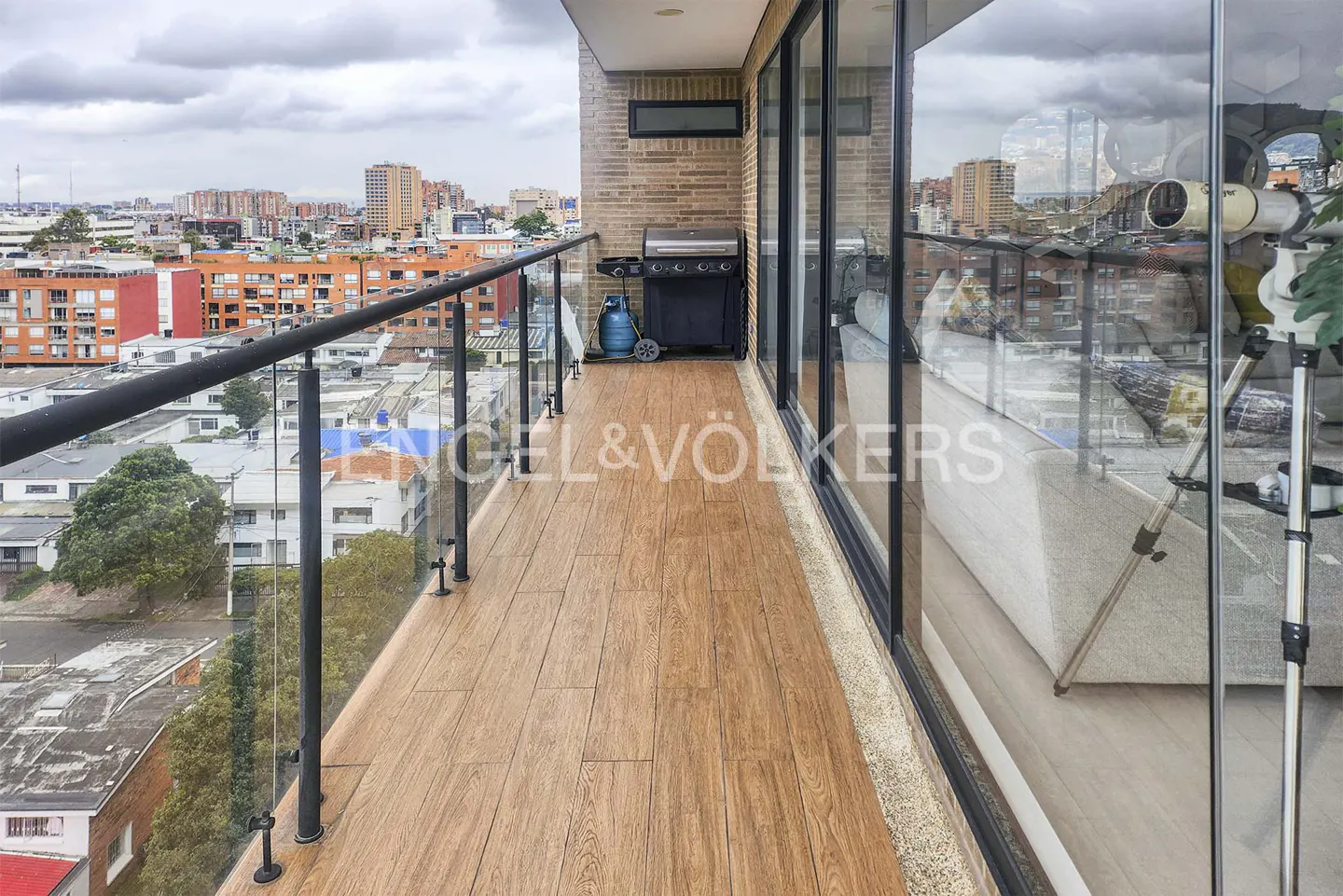 Modern balcony with wood-look tile, glass railing, and a grill, overlooking a city skyline under a cloudy sky.