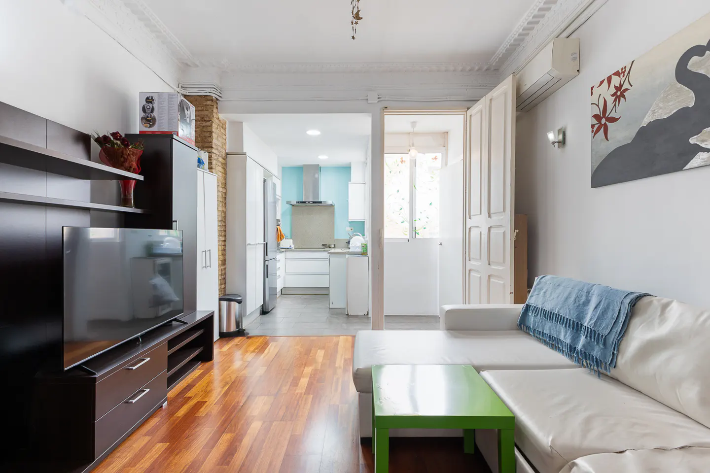 A bright living room with hardwood floors, a white sofa with a blue throw, and a green table. The kitchen is visible in the background.