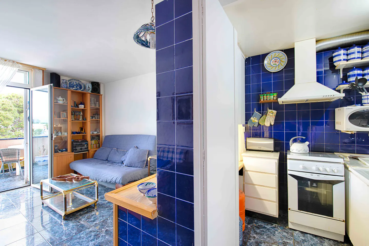 A view of a living room and kitchen with blue tile accents. The living room has a blue sofa, glass table, and balcony. The kitchen has white appliances.