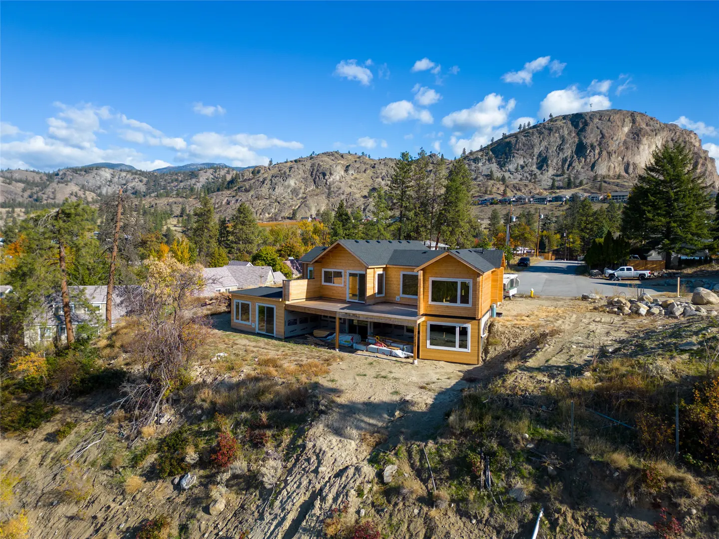 Two-story tan house with a deck, set on a hillside with a mountain backdrop under a blue sky.
