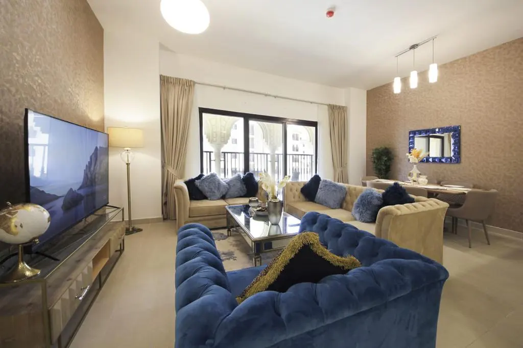 Living room with blue velvet chair, beige sofas, TV, and dining area. Neutral walls and curtains create a warm, inviting space.