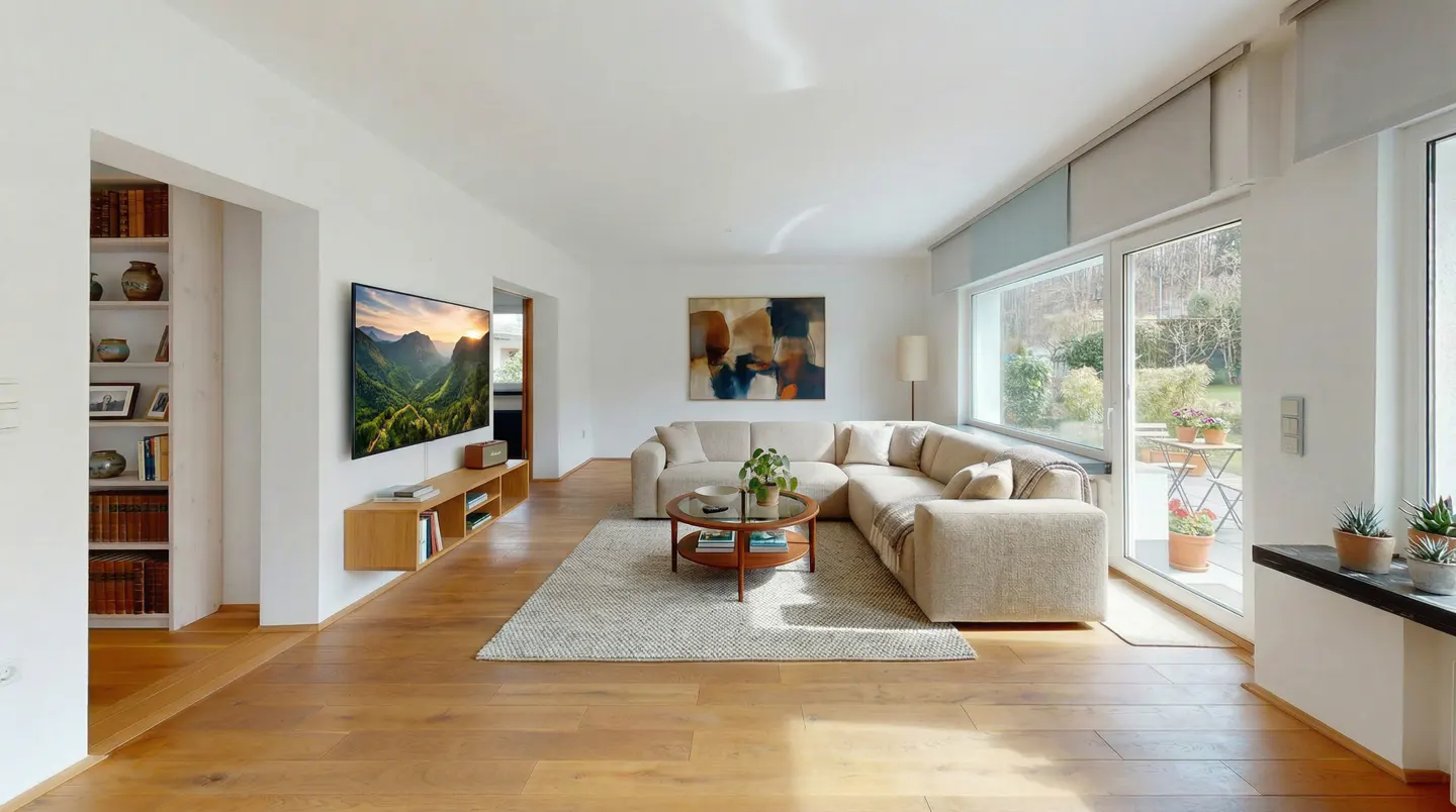 Bright living room with hardwood floors, a beige sectional sofa, and a large TV displaying a mountain landscape. A round coffee table sits on a patterned rug.