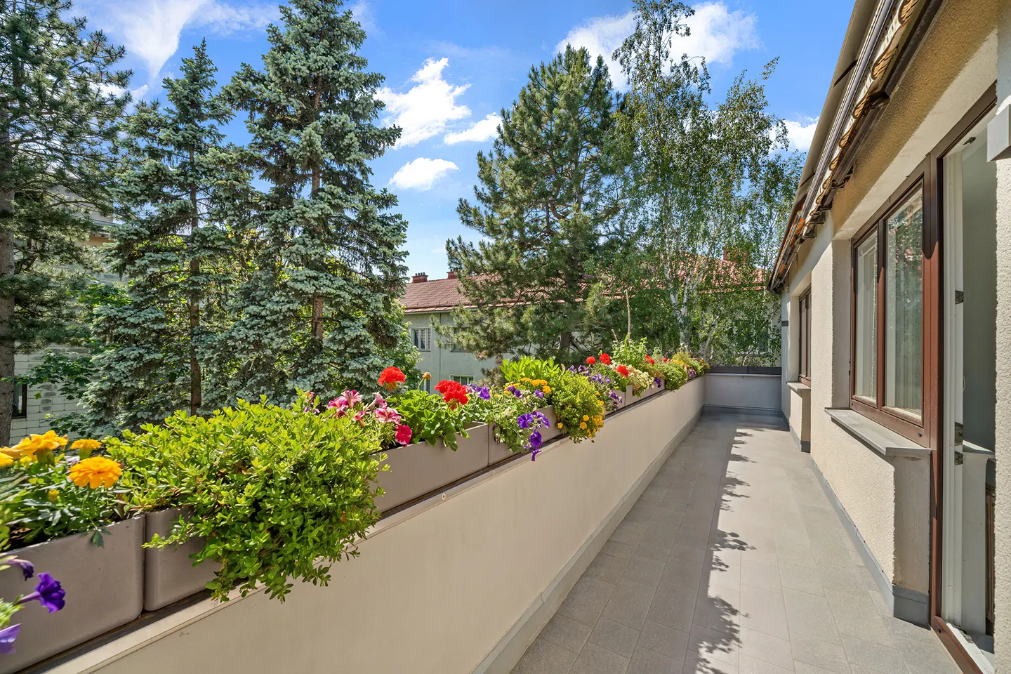 A sunny balcony with tiled flooring, featuring long planters filled with vibrant colorful flowers and lush green foliage.