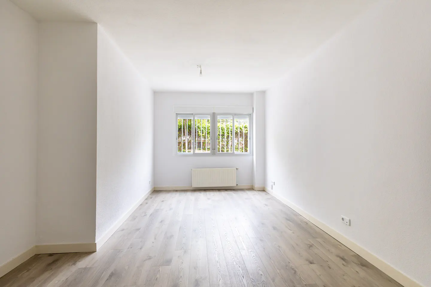 Bright, empty room with white walls and light gray wood floors. A window with white blinds looks out onto green trees. A white radiator sits below the window.
