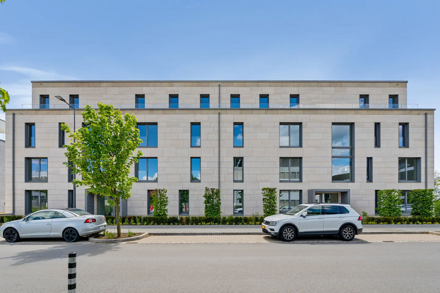 A three-story apartment building with a light stone facade and dark-framed windows, with two white cars parked in front.