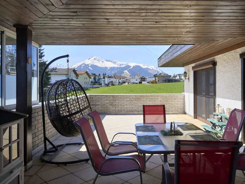 Covered patio with red chairs around a table, a hanging chair, and a view of snow-capped mountains.