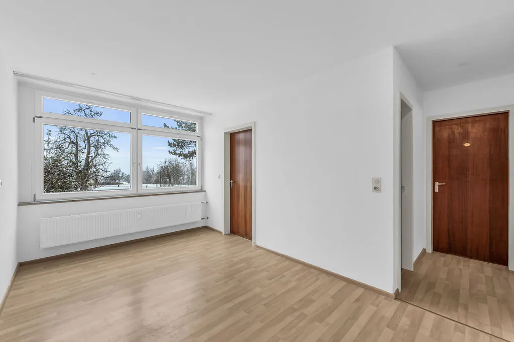 Bright, empty room with light wood floors, white walls, and a large window showing a snowy landscape. Two wooden doors are visible.