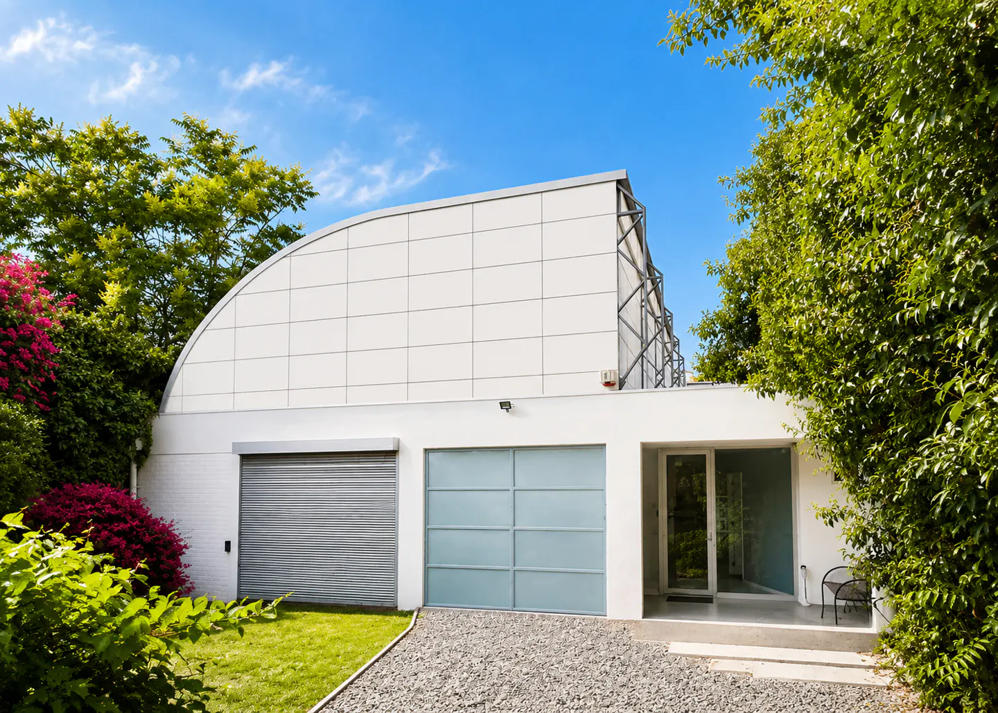 Modern white building with two garage doors, a glass door entrance, and a curved roof, surrounded by lush greenery.