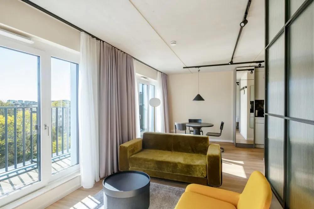 Bright living room with olive sofa, yellow chair, and round table. Balcony doors open to a view of trees and buildings.
