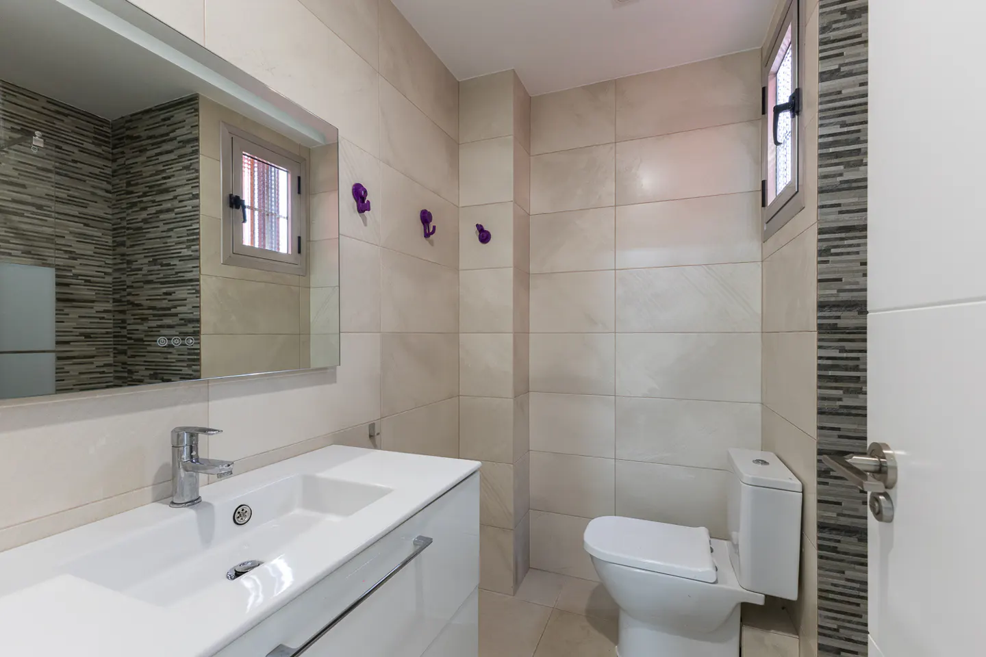 Modern bathroom with white vanity, chrome faucet, toilet, and beige tiled walls with decorative grey stone accents.
