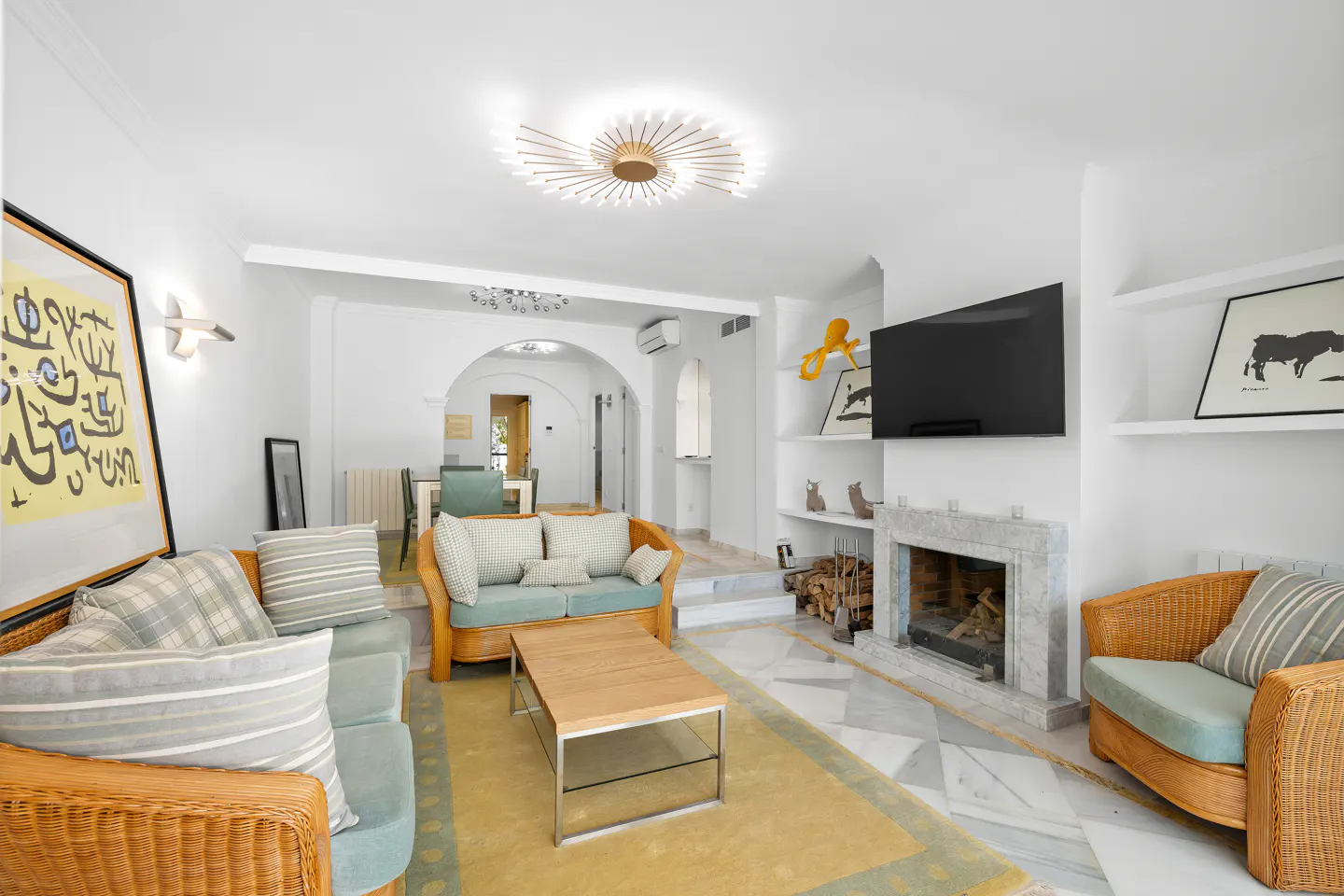Bright living room with wicker furniture, a marble fireplace, and a yellow rug on a marble floor. Artwork adorns the white walls.