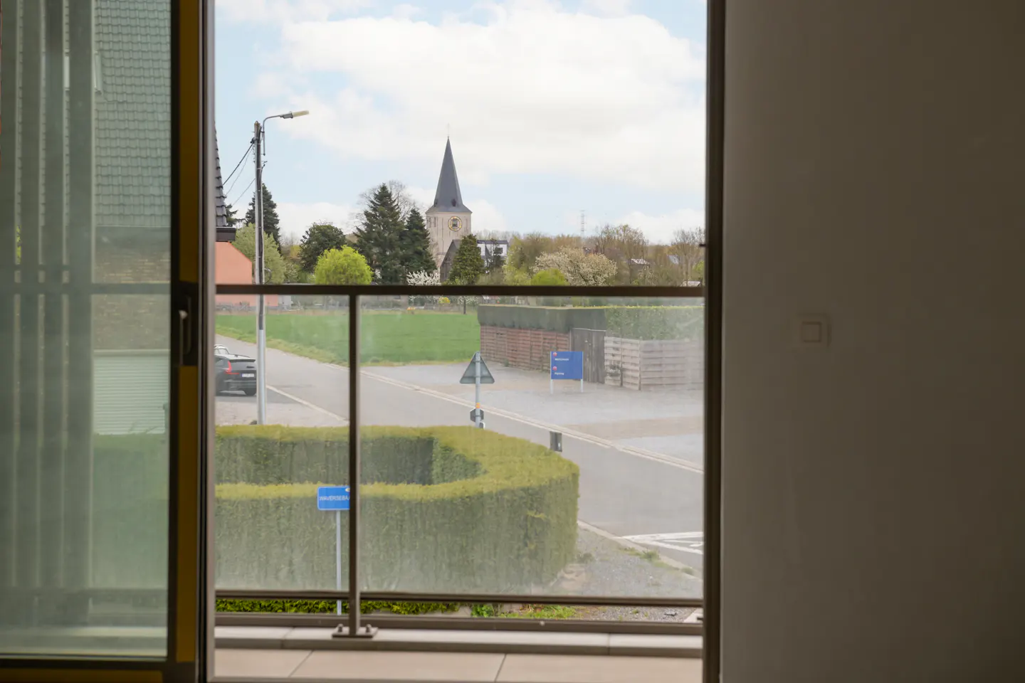 View from a balcony with glass railings overlooking a street, green hedges, and a church steeple in the distance.