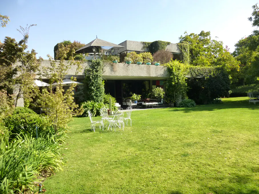 A modern concrete house with a green lawn, trees, and white outdoor furniture.