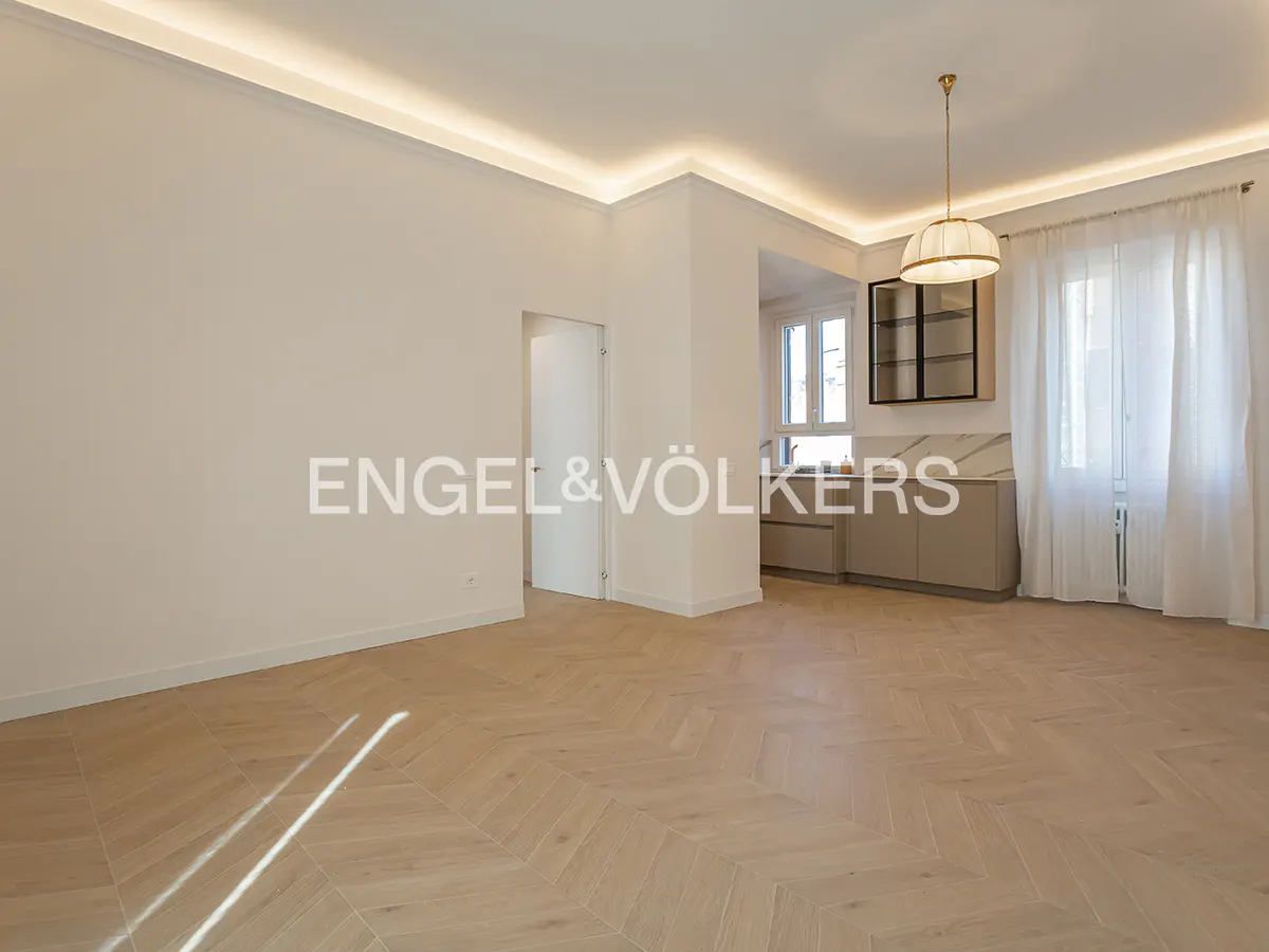 Bright, empty room with herringbone wood floors, white walls, and recessed lighting. Kitchenette visible through opening. Engel & Völkers logo on wall.