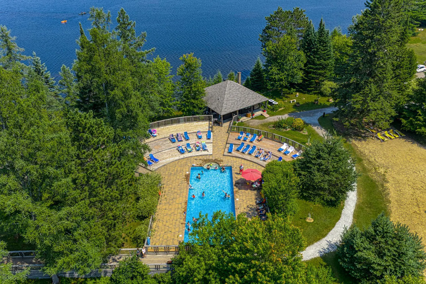 Aerial view of a blue pool surrounded by lounge chairs, a lake, and green trees.