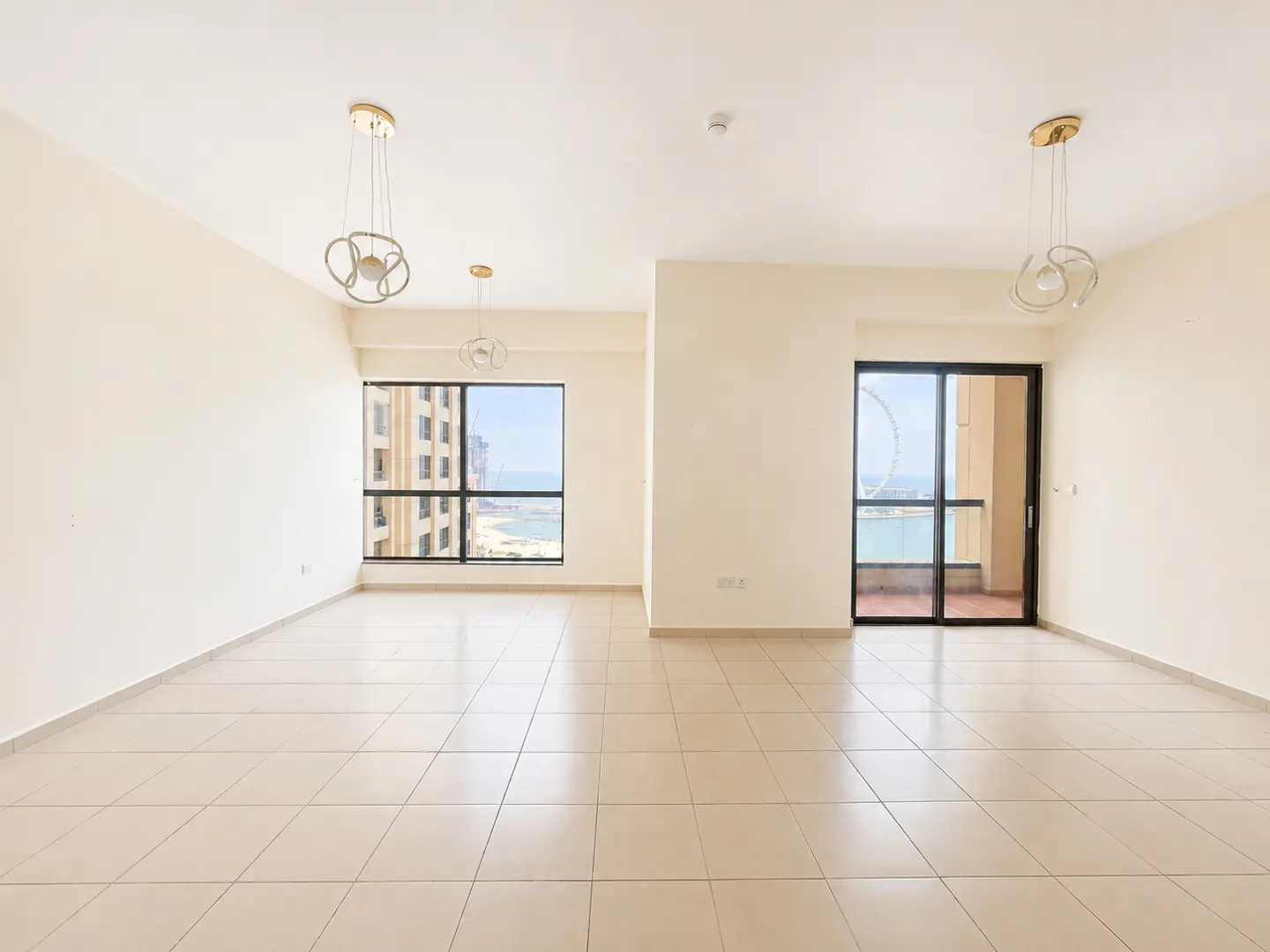 Empty, bright apartment with beige tile floors, white walls, and modern light fixtures. Windows offer ocean views. Balcony access via sliding glass doors.