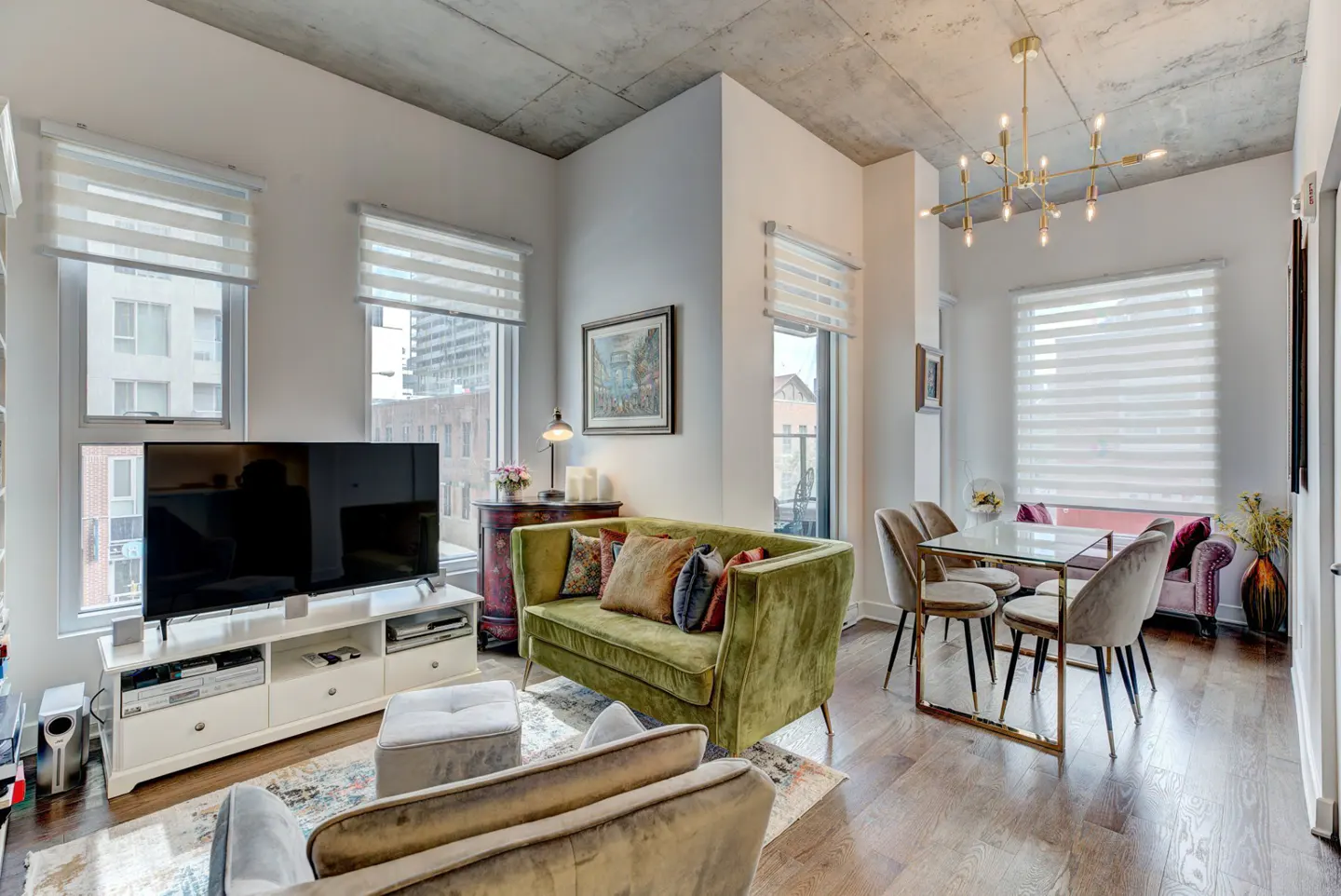 Bright living room with a green velvet sofa, TV, dining table, and large windows with white blinds. Concrete ceiling and hardwood floors.